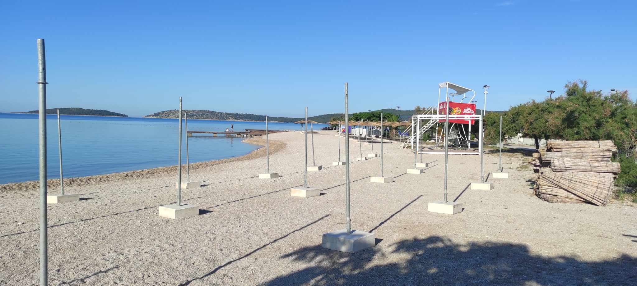 Plaža Rezalište s nizovima metalnih nosača suncobrana na betonskim postamentima, u pozadini spasilački toranj